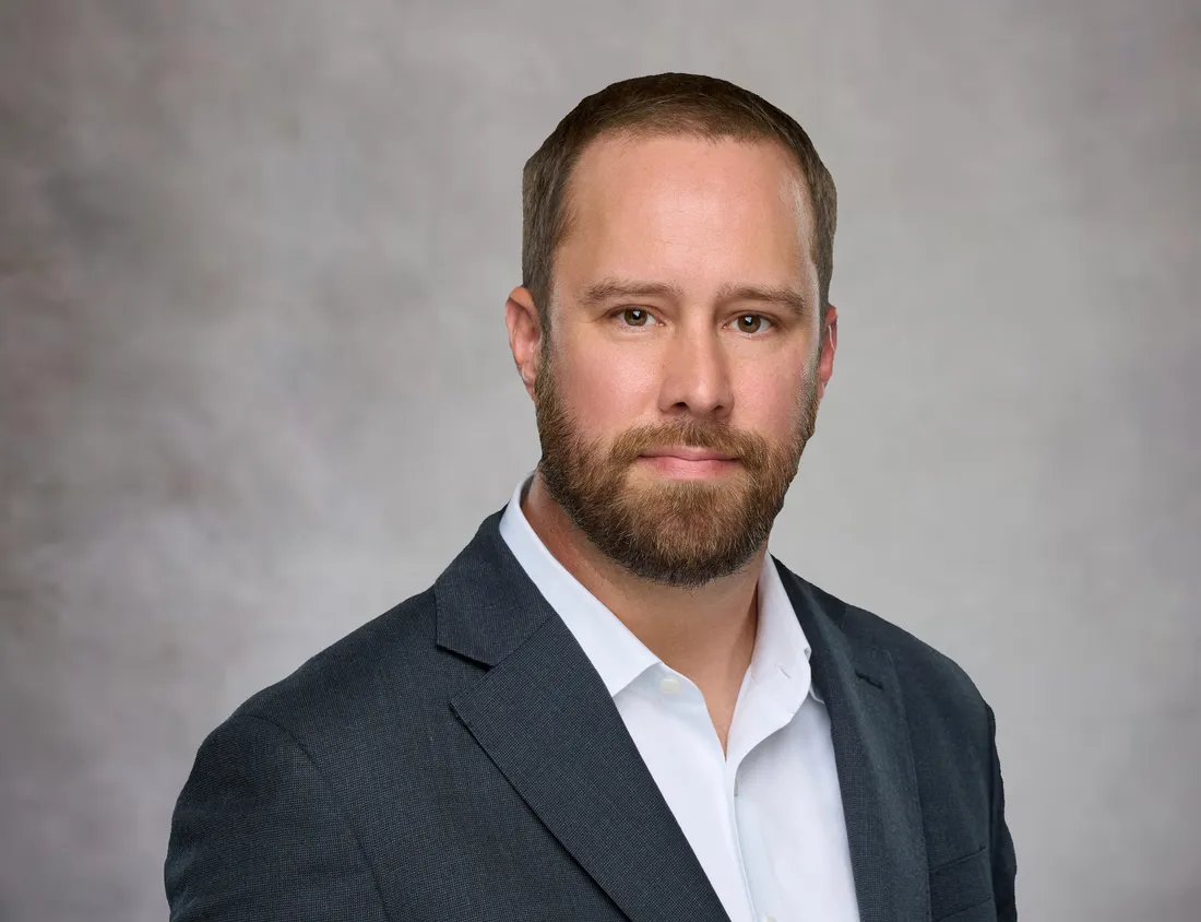 Ken Dickinson, wearing a charcoal blazer and open-collar white shirt, photographed against a warm gray backdrop.